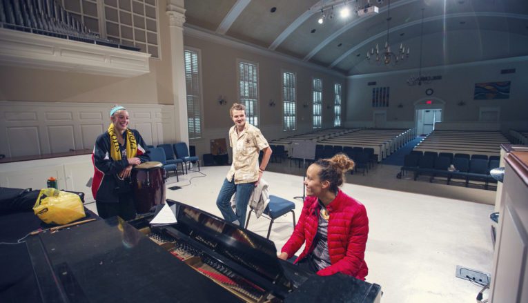 Three students on stage in auditorium, on sitting at piano